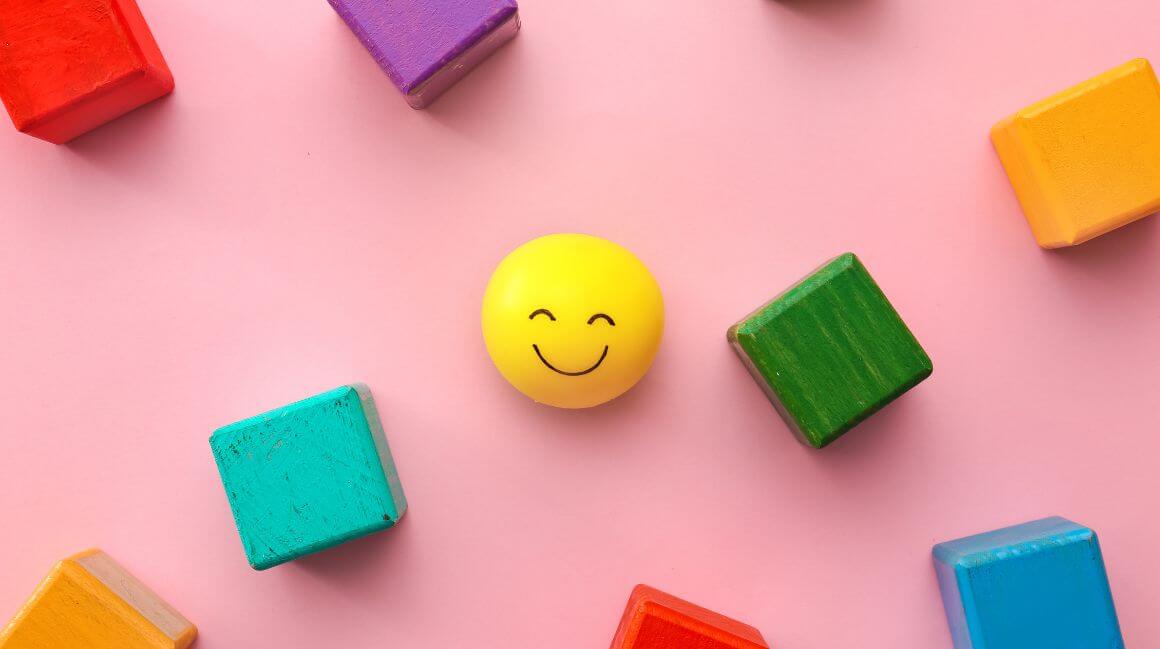 Ein lächelnder Smiley Bauklotz liegt neben anderen bunten Bauklötzen.