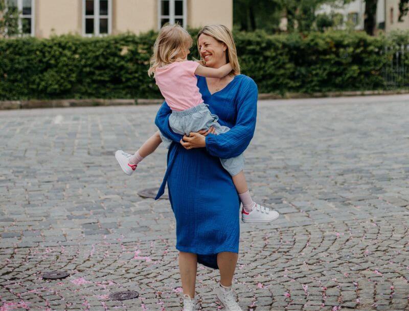 Blog_Headerbild_Sich_neu_erfinden_als_Schulkindeltern Eine Mutter hält ihr Schulkind auf dem Arm. Sie schauen sich in lächelnd an und wirken so, als ob sie sich glücklich im Kreis drehen.