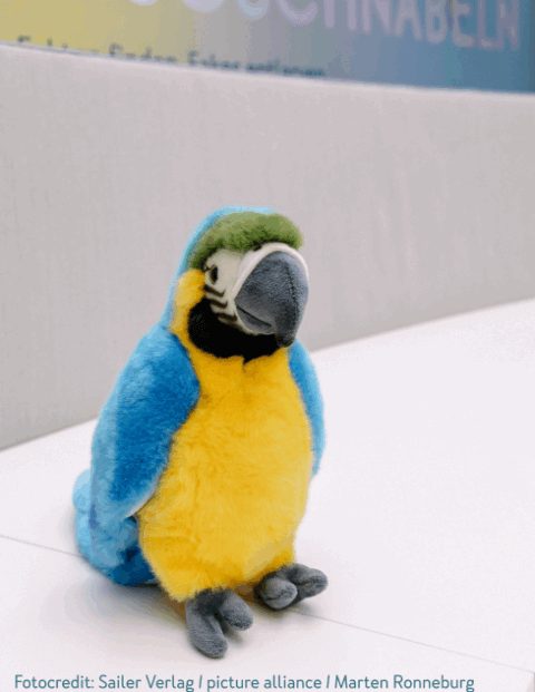 Ein Plüschpapagei sitzt auf einer weißen Oberfläche. Der Papagei hat ein blaues Gefieder, einen gelben Bauch und graue Füße. Im Hintergrund ist unscharf ein Abschnitt einer Wand mit Text zu sehen.