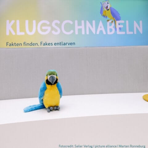 Ein Stoffpapagei sitzt auf einer weißen Oberfläche. Im Hintergrund ist ein Schild mit der Aufschrift 'KLUGSCHNABELN', darunter der Text 'Fakten finden, Fakes entlarven'. Ein gezeichneter Papagei ist ebenfalls auf dem Schild abgebildet. Fotocredit: Sailer Verlag / picture alliance / Marten Ronneburg.