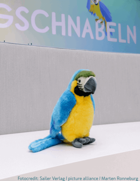 Ein Plüschpapagei mit blauem Gefieder und gelbem Bauch sitzt auf einer weißen Oberfläche. Im Hintergrund ist unscharf ein Teil eines Schriftzugs auf einer Leinwand zu sehen.