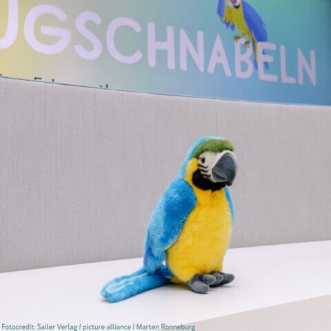 Ein Stoffpapagei in hellen Farben, hauptsächlich blau und gelb, sitzt auf einer weißen Oberfläche. Im Hintergrund ist ein Schild mit der teilweise sichtbaren Aufschrift 'Schnabeln' zu sehen.