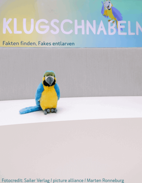 Ein Papageien-Plüschtier sitzt vor einer Hintergrundwand mit dem Schriftzug 'KLUGSCHNABELN' und dem Untertitel 'Fakten finden, Fakes entlarven'.