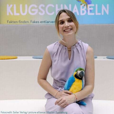 Eine lächelnde Frau sitzt vor einem Hintergrund mit der Aufschrift 'KLUGSCHNABELN - Fakten finden, Fakes entlarven'. Sie trägt ein lila ärmelloses Kleid und hält einen blauen und gelben Plüschpapagei. Einige bunte Kissen sind im Hintergrund auf Bänken platziert.