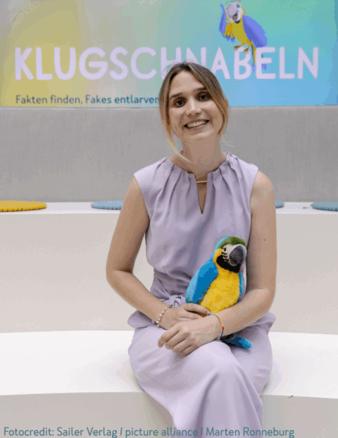 Eine lächelnde Frau in einem lila Kleid sitzt auf einer weißen Bank und hält ein Plüschtier eines blauen und gelben Papageis. Hinter ihr ist ein Poster mit der Aufschrift 'KLUCHSCHNABELN: Fakten finden, Fakes entlarven'.