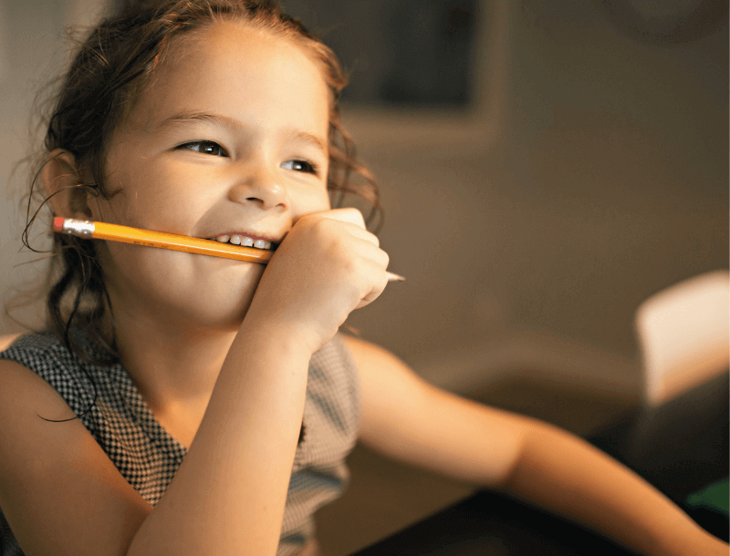 Blog_Headerbild_Lernplatz Ein Mädchen sitzt am Schreibtisch und hält einen Bleistift in der Hand. Denkt lächelnd über etwas nach.