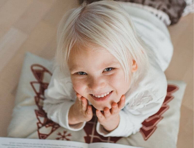 blog-headerbild-last-minute-geschenkideen Ein lächelndes Kind mit blondem Haar liegt auf einem Kissen und schaut freudig in die Kamera. Im Vordergrund ist ein aufgeschlagenes Buch oder Heft erkennbar.