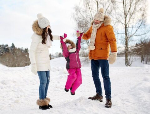 Ein lachendes Kind in rosa Winterkleidung wird von zwei Erwachsenen hochgehoben. Sie stehen im Schnee, alle tragen warme Winterkleidung mit Mützen und Handschuhen. Im Hintergrund sind schneebedeckte Bäume und ein klarer Himmel zu sehen.