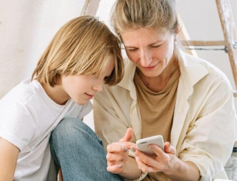 Eine erwachsene Person und ein Kind sitzen nahe beieinander und schauen gemeinsam auf ein Mobiltelefon. Die Szenerie wirkt harmonisch und interessiert. Beide tragen legere Kleidung.