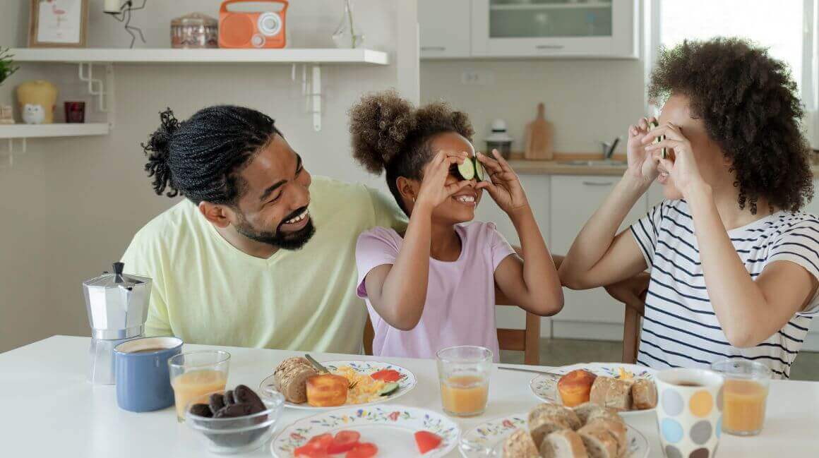 Eine Familie sitzt gemeinsam am Frühstückstisch in einer hellen Küche. Ein Mann, ein Kind und eine Frau lachen und das Kind hält Gurkenscheiben vor die Augen. Auf dem Tisch befinden sich Brötchen, Orangensaft, Kaffee und frisches Obst. Im Hintergrund sind Küchenutensilien und Dekorationen sichtbar.