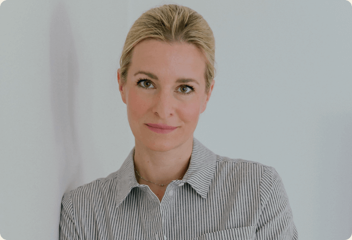 Eine Frau mit einem gestreiften Hemd steht vor einem neutralen Hintergrund und schaut freundlich in die Kamera.