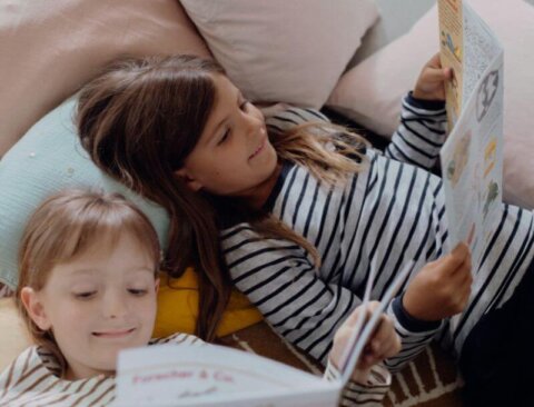 Zwei Kinder liegen nebeneinander auf Kissen und lesen Zeitschriften. Beide tragen gestreifte Oberteile und lächeln beim Lesen der Zeitschriften. Um sie herum befinden sich Kissen in verschiedenen Farben.