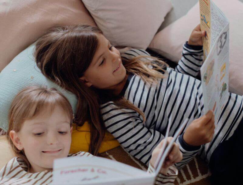 Blog_Headerbild_Lesemotivation-Kinder Zwei Kinder liegen nebeneinander auf Kissen und lesen Zeitschriften. Beide tragen gestreifte Oberteile und lächeln beim Lesen der Zeitschriften. Um sie herum befinden sich Kissen in verschiedenen Farben.