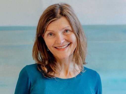 Eine Frau mit schulterlangem braunem Haar lächelt in die Kamera. Sie trägt ein blaues Oberteil. Der Hintergrund ist in hellen Blautönen gehalten. Chefredakteurin Gisela Lipsky