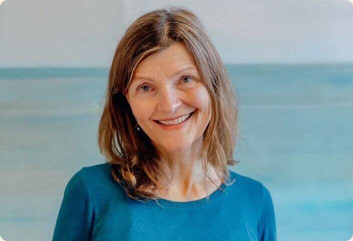 Eine Frau mit schulterlangem braunem Haar lächelt in die Kamera. Sie trägt ein blaues Oberteil. Der Hintergrund ist in hellen Blautönen gehalten. Chefredakteurin Gisela Lipsky