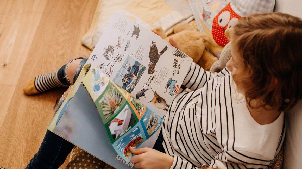 Ein Kind liegt auf dem Boden und liest eine illustrierte Zeitschrift. Das Kind trägt ein gestreiftes Oberteil und hat neben sich ein Kuscheltier. Auf der Zeitschrift sind Bilder von Tieren und Text zu sehen. Im Hintergrund ist ein Kissen mit Eulenmotiv erkennbar.
