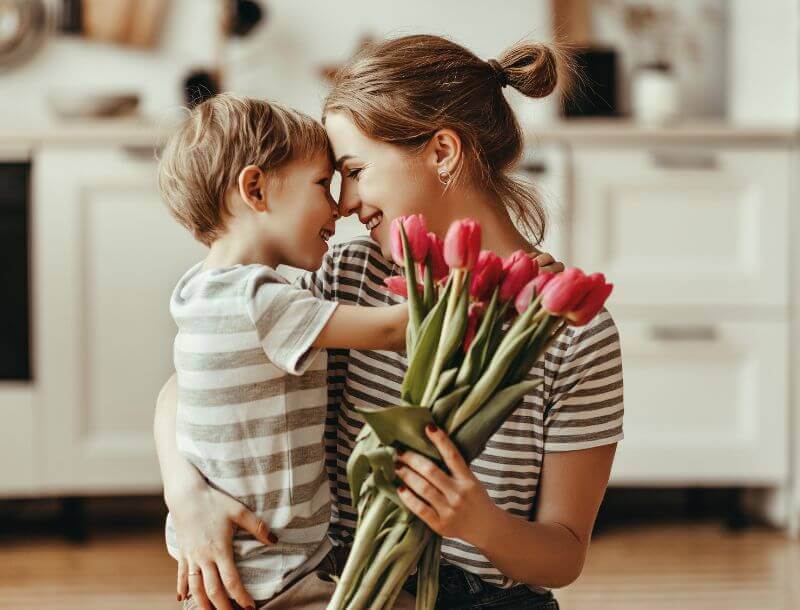 Blog_Headerbild_Gleichstellung Eine Frau und ein Kind in einem gestreiften T-Shirt umarmen sich herzlich in einer Küche. Die Frau hält einen Strauß mit rosa Tulpen. Beide lächeln und haben ihre Stirn aneinander gelegt.
