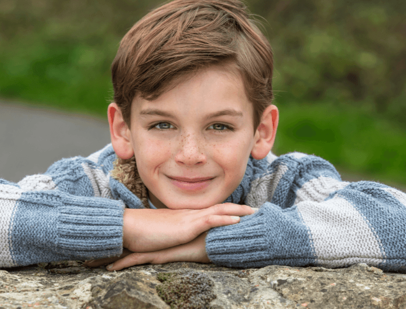 Blog_Fernandes-Ulmen Ein Junge mit braunen Haaren und Sommersprossen, der einen gestreiften Pullover trägt, liegt mit verschränkten Armen auf einem Felsen und lächelt in die Kamera. Im Hintergrund ist verschwommenes Grün zu sehen.