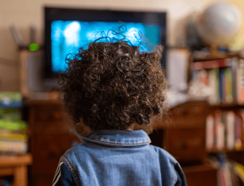 Ein Kind mit lockigem Haar, das vor einem Fernseher sitzt. Im Hintergrund sind unscharf ein Bücherregal und ein Globus zu sehen.