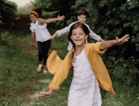 Drei Kinder laufen fröhlich auf einem grünen Waldweg. Das vordere Mädchen trägt ein weißes Kleid und eine gelbe Strickjacke. Die beiden anderen Kinder sind hinter ihr und ebenfalls fröhlich gestimmt. Der Hintergrund zeigt grüne Bäume und Büsche.