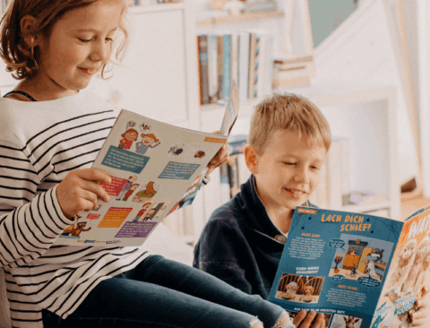 Zwei Kinder sitzen nebeneinander und lesen fröhlich in bunten Zeitschriften. Das Mädchen trägt ein gestreiftes Oberteil und das Junge ein dunkles Oberteil. Im Hintergrund ist ein Regal mit Büchern zu sehen.