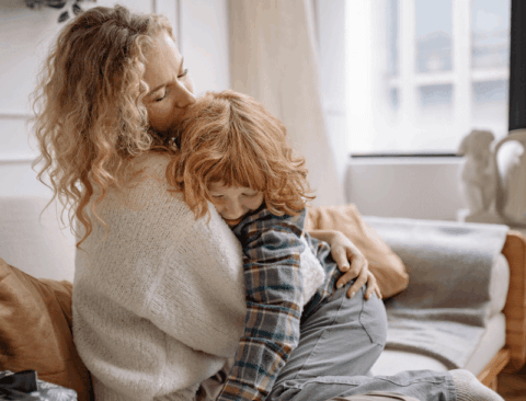 Eine Frau mit lockigem Haar sitzt auf einem Sofa und hält ein Kind mit rotem Haar im Arm. Beide wirken entspannt und liebevoll miteinander verbunden. Im Hintergrund sind ein großes Fenster und neutral gestaltete Wohnaccessoires zu sehen.