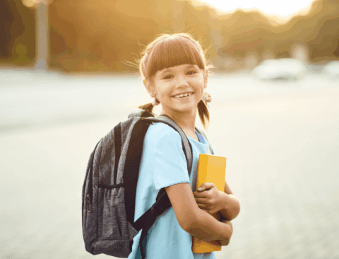 Ein lächelndes Mädchen mit braunen Haaren und Pferdeschwanz, das einen grauen Rucksack trägt und ein gelbes Buch hält. Sie steht im Freien bei sonnigem Wetter.