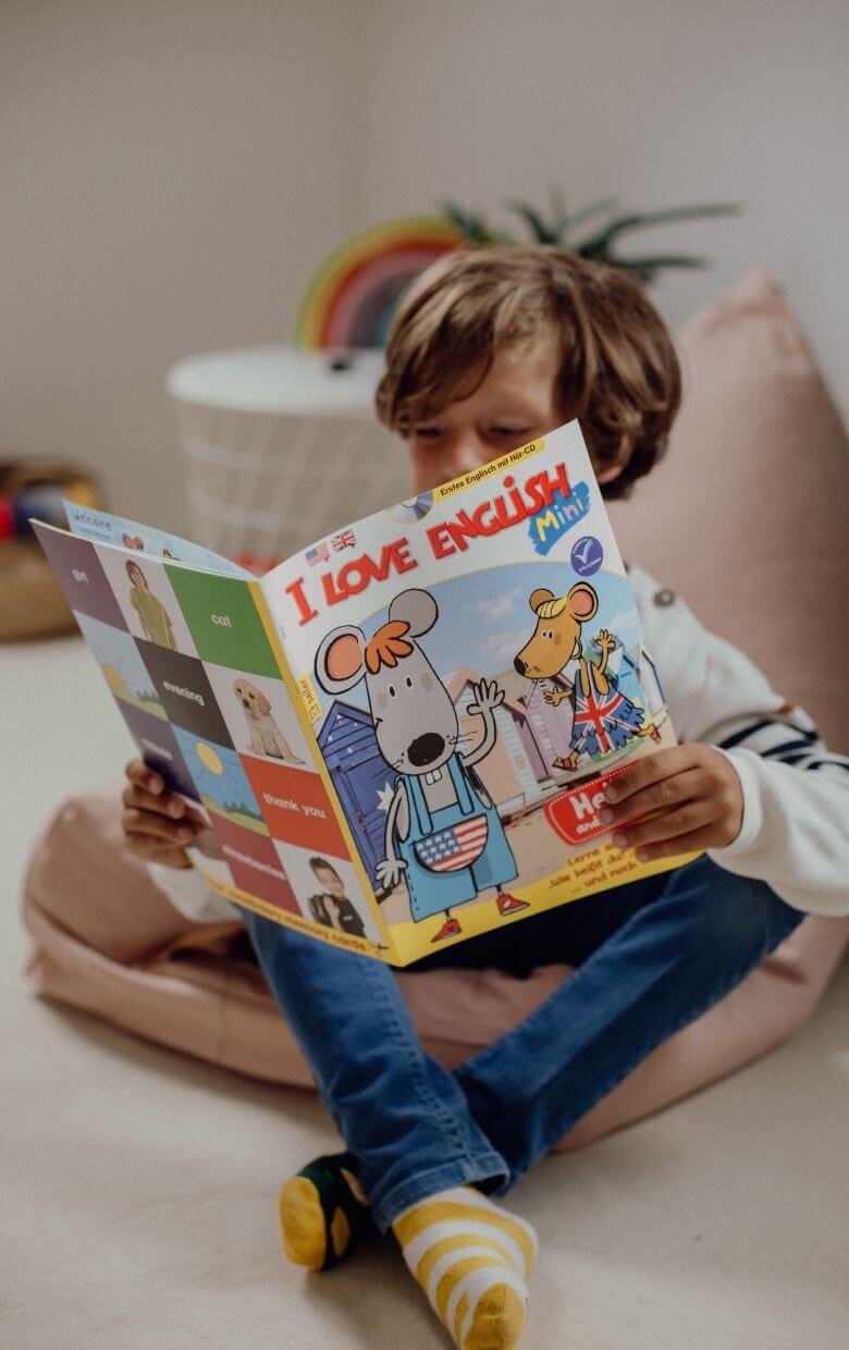 Ein Kind sitzt auf einem rosa Kissen und liest ein buntes Kinderheft mit dem Titel 'I Love English Mini'. Auf dem Cover sind zwei zeichentrickartige Figuren, eine Maus und ein Hund, zu sehen, die vor einer Kulisse mit Strandhütten stehen. Das Kind trägt blaue Jeans und gestreifte Socken.