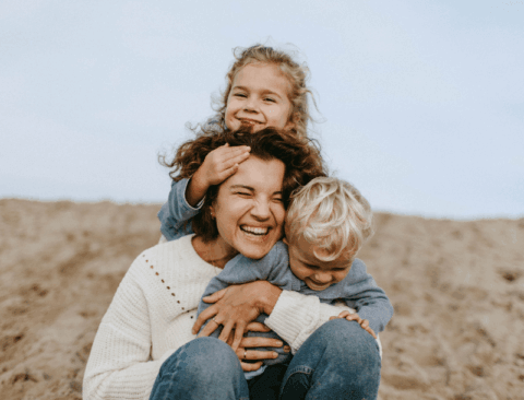 Eine Frau umarmt zwei Kinder und lächelt fröhlich an einem Sandstrand. Eines der Kinder sitzt auf ihren Schultern, während das andere in ihrem Schoß kuschelt. Sie tragen warme Kleidung und scheinen einen schönen Moment im Freien zu genießen.