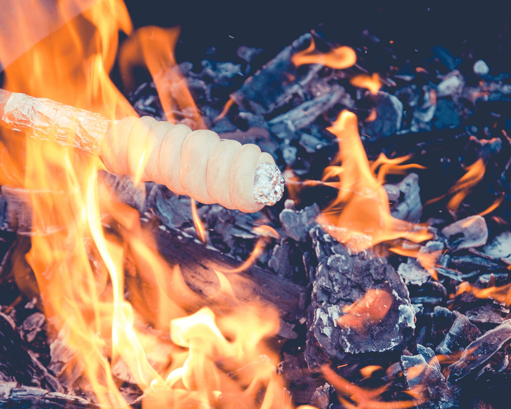 Ein Stockbrot wird über einem offenen Feuer gebacken. Der Teig ist spiralförmig um einen Stock gewickelt, der teilweise mit Aluminiumfolie bedeckt ist. Flammen und glühende Kohlen sind im Hintergrund sichtbar.
