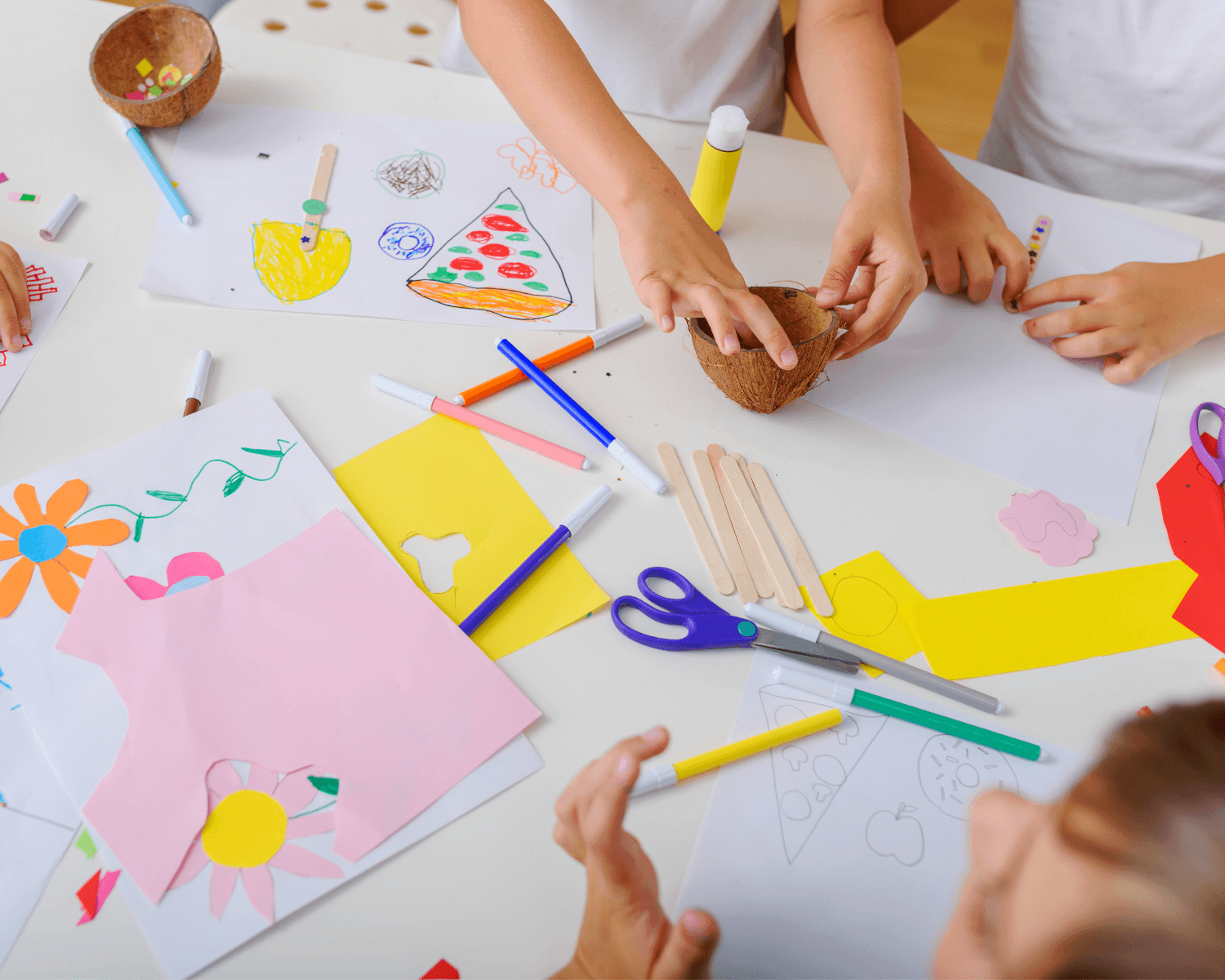 Kinder basteln an einem Tisch mit verschiedenen Bastelmaterialien wie Buntstiften, Scheren, Papierschnipseln und Eisstielen. Auf dem Tisch sind Zeichnungen mit Blumen und einer Pizza zu sehen.